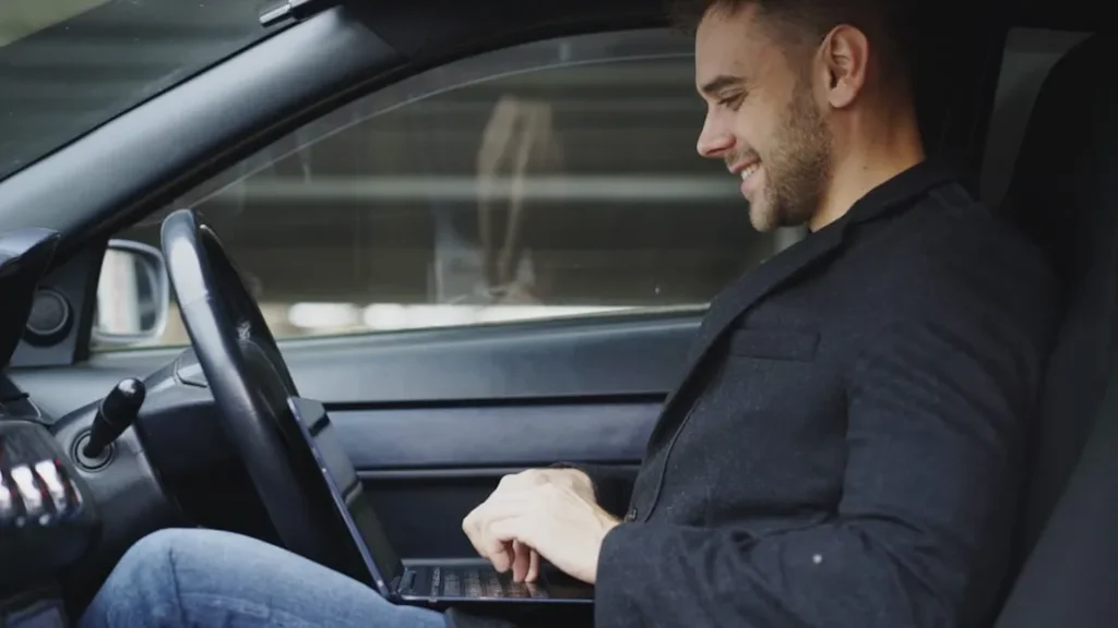 A smiling man in a black jacket and blue jeans sits in the driver's seat of a car, typing on a laptop resting on his lap.