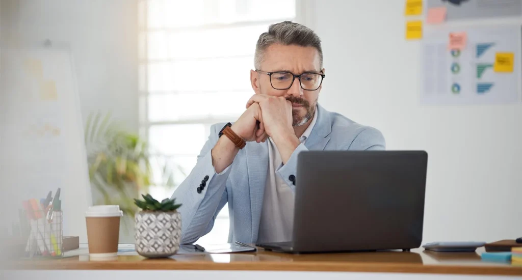 A bearded man with glasses sits at a desk, hands under chin, focused on a laptop in an office with a whiteboard and papers.