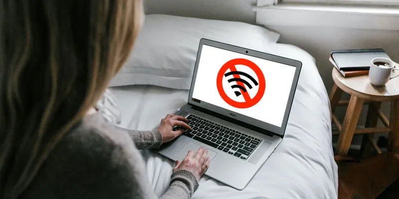 A person on a bed uses a laptop with a "no Wi-Fi" symbol on the screen, indicating a lack of internet connection.